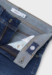 Μπλε τζιν παντελόνι με αντίθετες κίτρινες ραφές, μεταλλικό φερμουάρ και κλείσιμο με κουμπί. Το εσωτερικό διαθέτει ανοιχτό ύφασμα με υποβαθμισμένο σχέδιο.