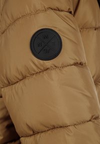 Rudas pūkuotasis žieminis švarkas su lygiu, blizgiu paviršiumi, ant rankovės esantis apvalus juodas guminis logotipo ženkliukas, pažymėtas "NMT" ir "97".
