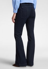 Tmavě modré džíny s rozšířenými nohavicemi a středním pasem, s dvěma zadními kapsami a jemnými švy, nošeny s světle modrou košilí.