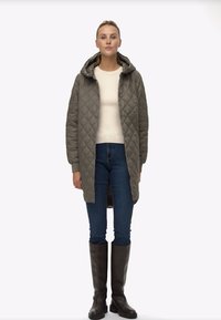 Vooderdatud oliiviroheline pikk mantel kapuutsiga, millele on lisatud hele sviiter, sinised teksad ja põlvikud pruunid saapad. Sile tekstuur, minimaalne disain.