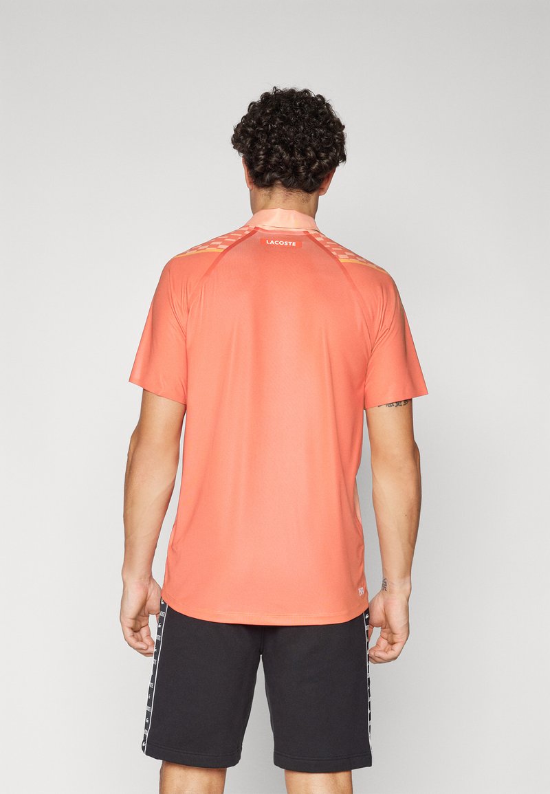 Korallefarbendes Sport-T-Shirt mit glatter Textur, reflektierenden Akzenten und Lacoste-Branding auf der Rückseite; getragen mit schwarzen Shorts mit seitlichen Streifen.