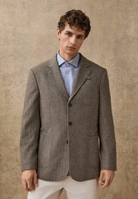 Brown herringbone-patterned blazer with two front pockets, notched lapels, and a single-breasted design, paired with a light blue shirt.