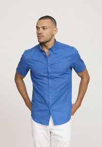 Chemise à manches courtes, boutonnée, en tissu bleu vif avec une texture lisse, dotée d'un col et d'un détail de logo discret sur la poitrine.