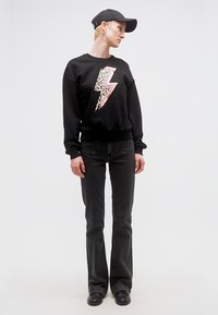 Άτομο που φοράει μαύρο πουλόβερ με αστραπή σε σχέδιο λεοπάρ, μαύρα καμπάνα τζιν, μαύρο καπέλο και μαύρα παπούτσια, εκείνο να στέκεται μπροστά σε λευκό φόντο.