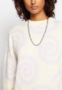 Cremfarbener Pullover mit lavendelfarbenen Wirbelmustern, strukturiertem Stoff und geripptem Ausschnitt. Enthält eine silberne Kettenhalskette.