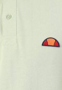 Šviesiai žalia polo marškinėlių su tekstūruota medžiaga, kuriuose kairėje krūtinės pusėje yra mažas, daugiakampis logotipas (oranžinės ir raudonos spalvos).