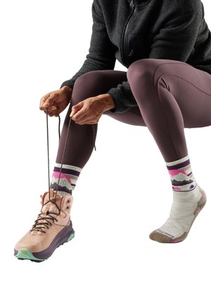 Person wearing purple leggings and mountain-patterned socks tying lace of pink hiking boot on one foot.