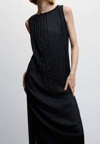 Vestido negro sin mangas con una tela texturizada y pliegues verticales, cuello alto y una abertura lateral para mayor movimiento.