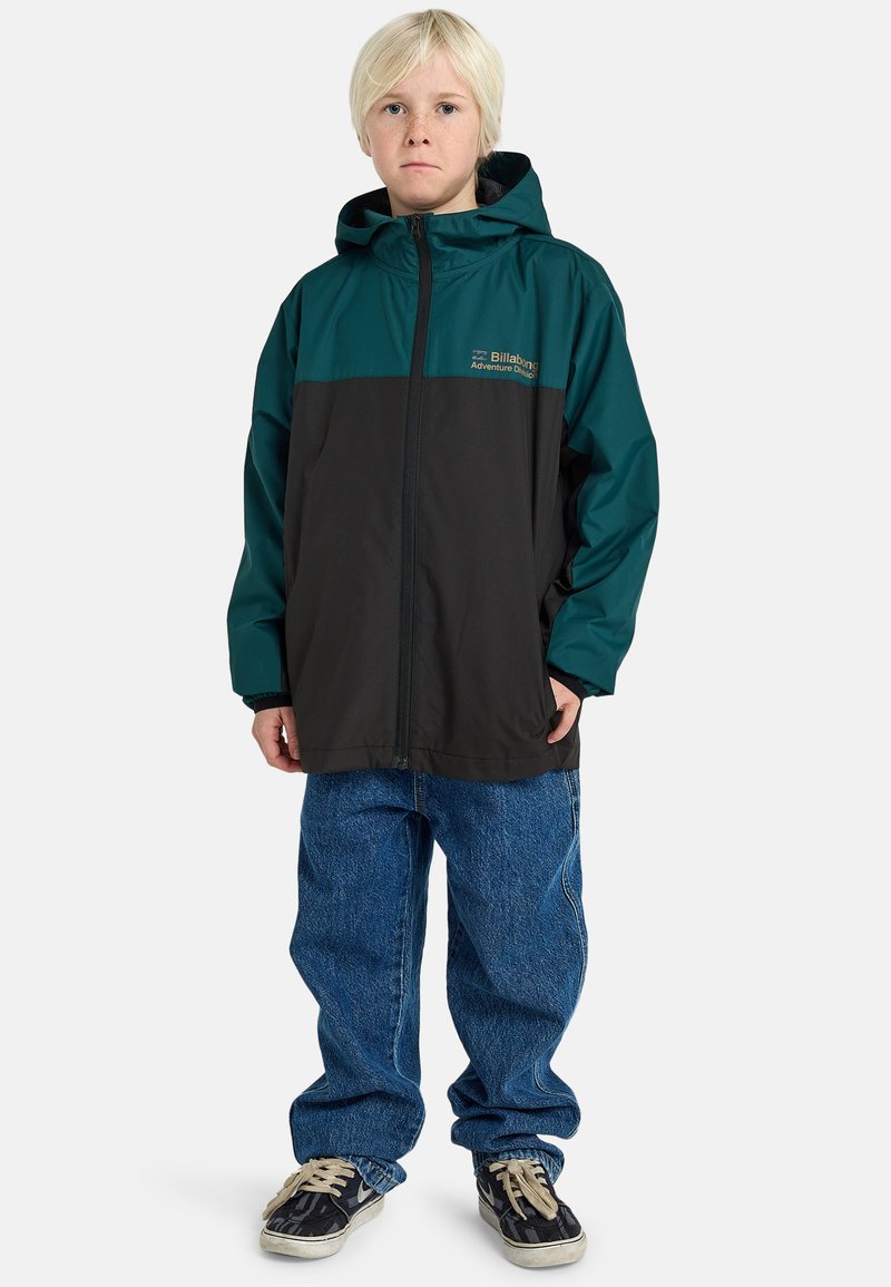 Teal und schwarze Windbreaker-Jacke mit Reißverschluss, Kapuze und Billabong-Logo. Kombiniert mit weit geschnittenen blauen Jeans und schwarzen Sneakers.