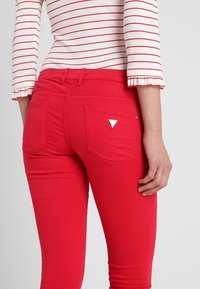 Jeans skinny rouges à finition texturée, avec deux poches arrière et un petit accent métallique triangulaire près de la poche droite.