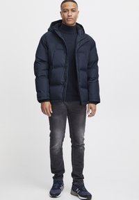 Jachetă puffer bleumarin cu glugă; design matlasat; închidere cu fermoar și capse în față. Asortată cu blugi închiși la culoare și pantofi sport albastre.