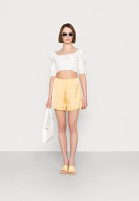 Top cropped branco com detalhes de renda plissada, calções amarelos de cetim, sandálias amarelas de deslizar e uma mala branca, contra um fundo simples.