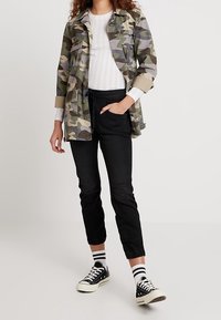 Kamikaze-mönstrad jacka med uppvikta ärmar, vit ribbad topp, svarta byxor och svarta sneakers med vita sulor samt randiga strumpor.