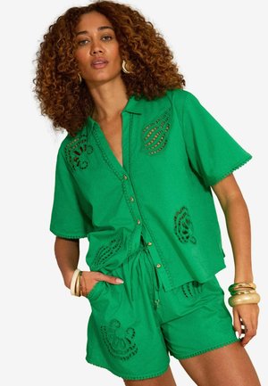 Lokkis juustega naine, kes kannab rohelist lühikeste varrukatega nööbitavat särki ja sobivaid lühikesi pükse lõigetega mustritega ning kuldseid käevõrusid.