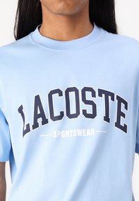 Světle modré bavlněné tričko s výrazným námořnickým textem, který uvádí "LACOSTE" a "SPORTSWEAR", s kulatým výstřihem, krátkými rukávy a hladkým povrchem.