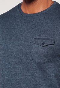 Detailní záběr osoby v modrém texturovaném tričku s kulat�ým výstřihem a knoflíkovou náprsní kapsou na levé straně.