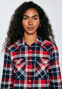 Red and navy plaid flannel shirt with two chest pockets, snap buttons, and a classic collar. Soft texture and long sleeves.
