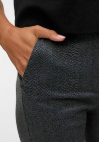 Pantalon à motifs en chevrons gris avec une poche latérale. La vue en gros plan montre la texture du tissu et une main reposant dans la poche.