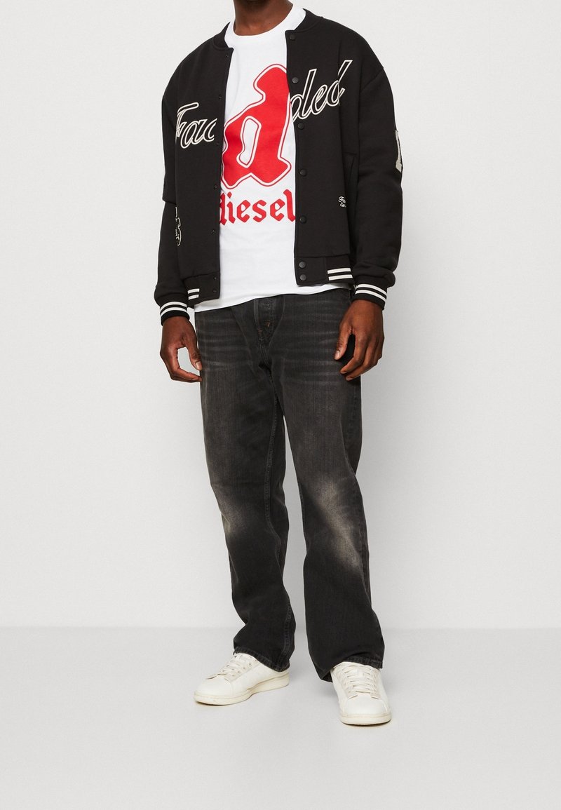 Svart varsityjacka med vita detaljer, röd grafisk t-shirt, mörka jeans och vita sneakers. Jackan har broderade detaljer.