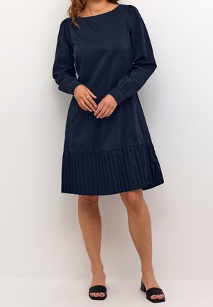 Nainen, jolla on yllään tummansininen pitkähihaisen mekon laskostettu helma, seisoo kädet ristissä ja jalassaan mustat avoimilla varpailla varustetut korkokengät.