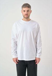 Weißes Langarm-T-Shirt aus Baumwolle mit Rundhalsausschnitt und Grafiken auf den Ärmeln. Kombiniert mit schwarzen Hosen.
