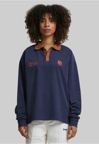 Marineblå langermet polo med brun krage og knapper. Har brodert tekst: "Berlin & New York" og logo på brystet.