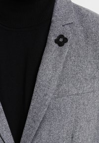 Szary wełniany blezer o fakturowanej tkaninie; posiada czarną przypinkę w kształcie kwiatu na klapie oraz czarny golf pod spodem.