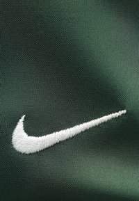 Το λογότυπο της Nike κεντημένο με άσπρες κλωστές σε υφασμάτινο φόντο σκούρου πράσινου χρώματος με υφή, που διαθέτει διακριτική λάμψη και σφιχτό ράψιμο για αντοχή.