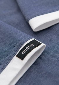 Modrá tkanina s texturovanou vazbou a bílým lemem, s černou etiketou s nápisem "ombre" v bílé barvě. Zaměření na detail rukávu.