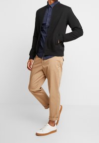 Svart zip-up bomberjacka med ribbat mönster, kombinerad med en mörkblå mönstrad skjorta och ljusbeige chinos, samt vita sneakers med bruna sulor.