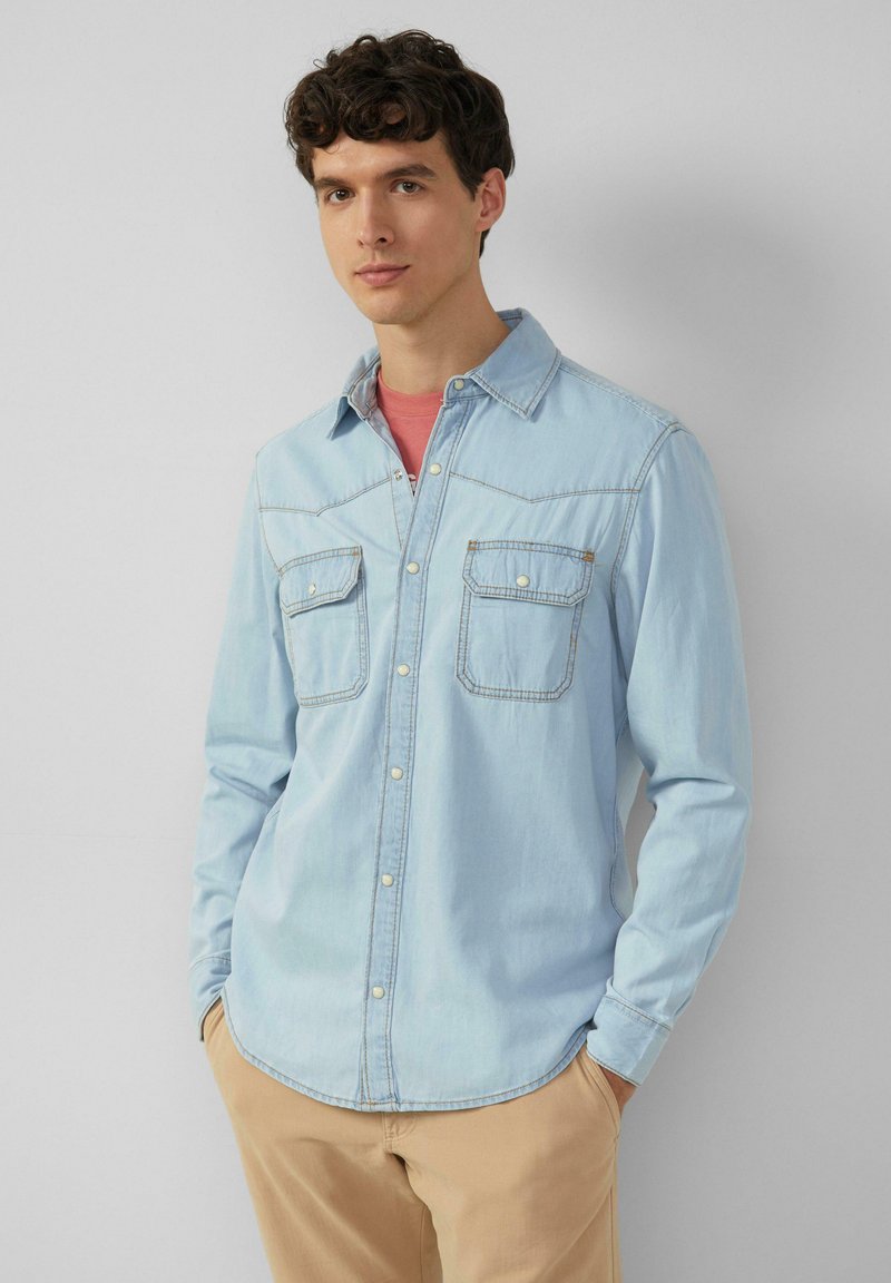 Camicia di jeans blu chiaro con due tasche sul petto, chiusura a bottoni e orlo curvo. Presenta cuciture a contrasto e un design con colletto classico.