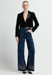 Blazer recortado de terciopelo negro con escote en V profundo y solapas de satén, combinado con jeans de cintura alta azul oscuro con adornos metálicos.