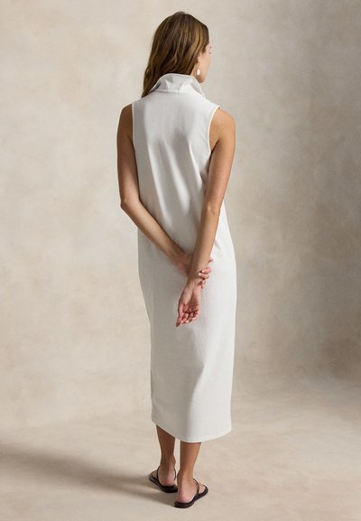 Femme debout, dos à la caméra, portant une robe midi blanche sans manches et des sandales plates noires sur un fond beige uni.