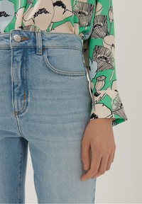 Blusa verde a stampa floreale con sfondo crema, abbinata a jeans a vita alta blu chiaro con il classico design a cinque tasche.