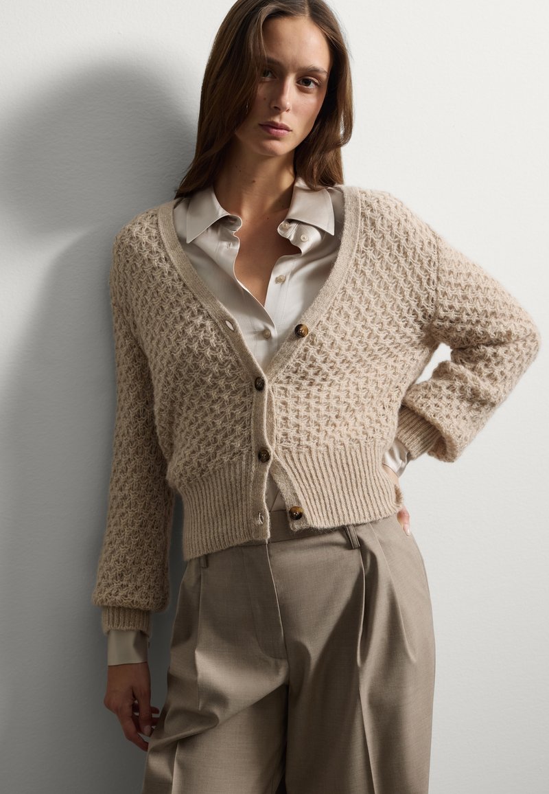 Cardigan beige testurizzato con scollo a V e chiusure a bottoni marroni, abbinato a una camicia chiara con bottoni e pantaloni beige larghi.
