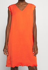 Oranje mouwloos jurkje met een V-hals, zachte textuur en gegolfde zoom. Lichtgewicht stof met een relaxte pasvorm.
