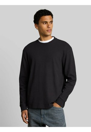 Junger Mann mit kurzen lockigen Haaren, der ein schwarzes Langarmshirt über einem weißen Unterhemd und blauen Jeans trägt, die Hand in der Tasche, schaut nach rechts.