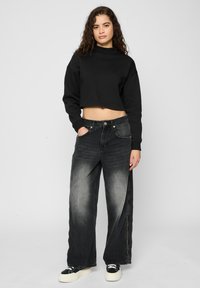 Sort kort genser med lange ermer, kombinert med løstsittende svarte jeans med falmet tekstur og sidelommer med glidelås, komplettert med svarte joggesko.