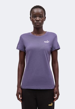 Γυναίκα με μωβ μπλουζάκι Puma και μαύρο παντελόνι Puma στέκεται μπροστά σε απλό ανοιχτό φόντο.