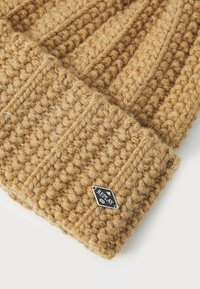 Gebreide beanie in een warme, lichtbruine kleur met een chunky textuur en ribbelontwerp, voorzien van een klein zwart en zilveren merketiket.