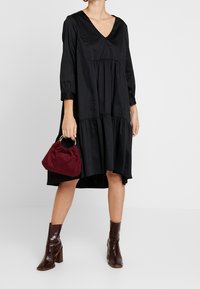 Schwarzes, gestuftes Kleid mit tiefem V-Ausschnitt und drei Viertelärmeln. Kombiniert mit einer burgunderfarbenen Wildleder-Handtasche und braunen Ankle-Boots mit viereckigen Absätzen.
