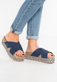 Fötter i blå denim krisscross plattformsandaler med vävda beige och marinblå sulor, som är matchade med uppvikta ljusblå jeans.