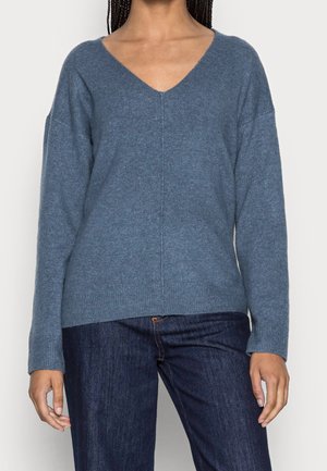 Frau trägt einen blauen V-Ausschnitt Pullover mit langen Ärmeln und dunkelblaue Jeans, steht vor einem schlichten hellen Hintergrund.