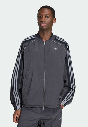 Young man standing with right hand in pocket wearing a dark gray Adidas tracksuit with three light gray stripes on sleeves.