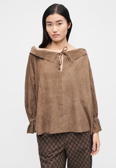 Ung kvinne iført en løs brun semsket topp med bred krage knyttet foran og mønstrete brune bukser, stående foran en ensfarget bakgrunn.