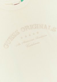 Cremfarbener Sweater aus strukturiertem Stoff, mit geprägtem "GUESS ORIGINALS"-Text und Sternen in einem zentralen Design.