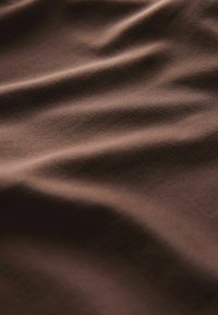 Tissu en coton marron avec une texture lisse, présentant de légères rides et une couleur uniforme, réfléchissant la lumière de manière douce et atténuée.