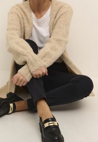 Beige gestrickte Strickjacke mit gerippten Bündchen, weißes T-Shirt, schwarze Jeans und schwarze Mokassins mit goldenen Akzenten. Lässige Sitzposition.
