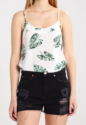 Mujer vistiendo una camiseta sin mangas blanca con estampados de hojas verdes y pantalones cortos vaqueros negros desgastados con bajos deshilachados.