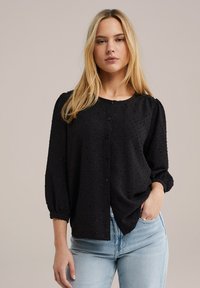 Blusa nera in tessuto strutturato, con bottoni frontali, scollatura rotonda e maniche a corno di tre quarti, abbinata a jeans in denim azzurro chiaro.